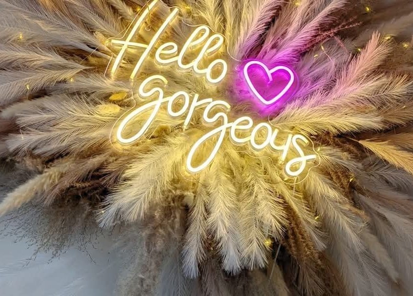 Illuminated 'Hello Gorgeous' sign at Bespoke Brows & Lashes, Havant, England, GB with decorative pampas grass.