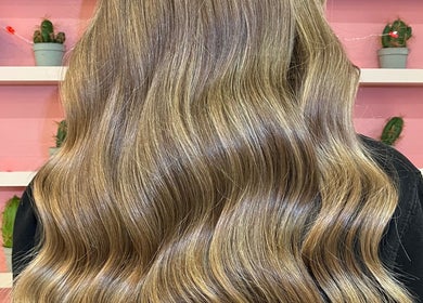 Long, wavy blonde hair styled at JessicaLibbyHair in Hinckley, England, GB. Background features pink wall and cacti.