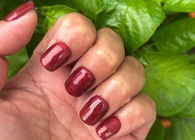 Crimson glitter nails expertly done at Trulyou, Swindon, England, GB, amidst vibrant green leaves.