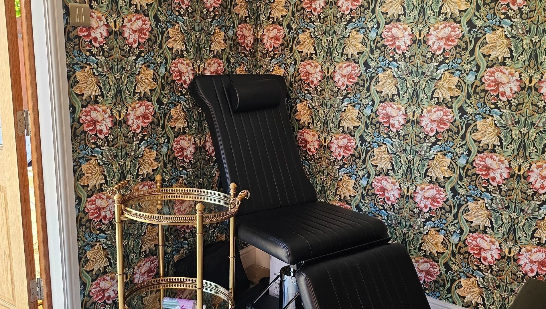 Elegant black chair in Monique's Hair Extension Boutique, Great Finborough, England, with floral wallpaper.