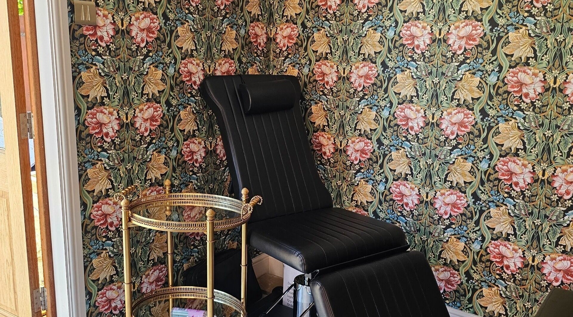 Elegant black chair in Monique's Hair Extension Boutique, Great Finborough, England, with floral wallpaper.