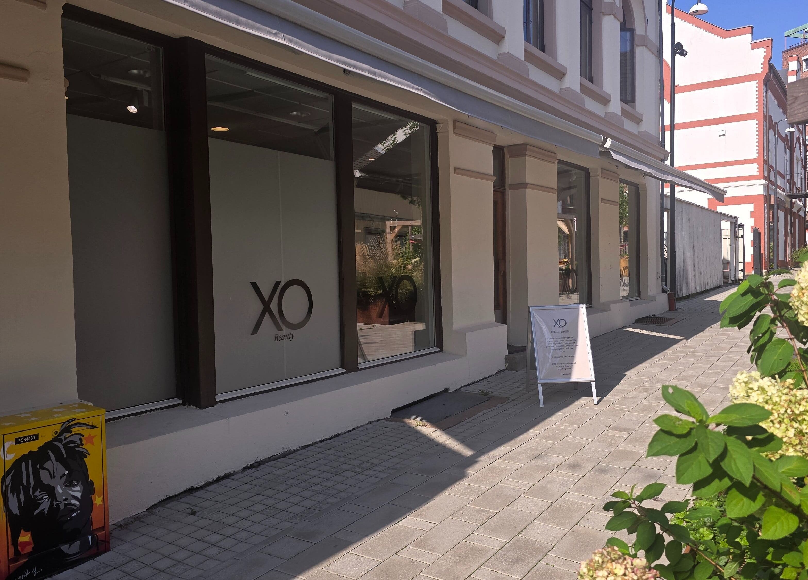 Utendørs fasade av XO Beauty & Academy AS, Sarpsborg, Østfold, NO, med blomster i forgrunnen.