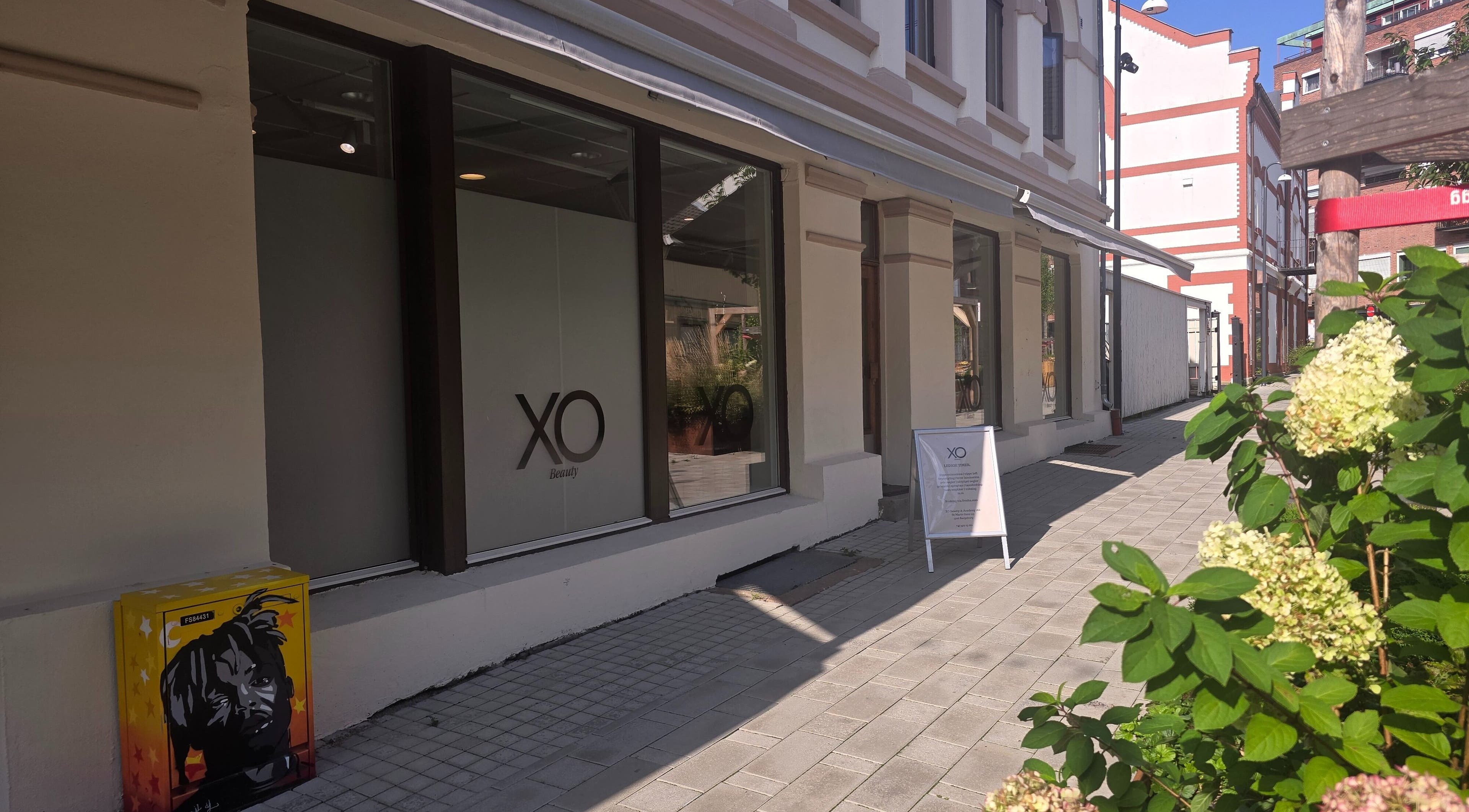 Utendørs fasade av XO Beauty & Academy AS, Sarpsborg, Østfold, NO, med blomster i forgrunnen.