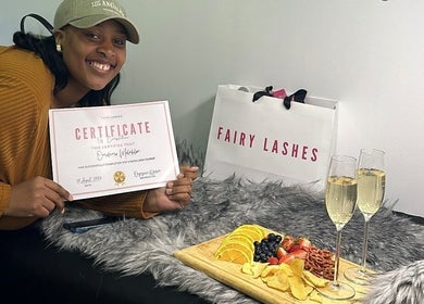 Smiling person at Fairy Lashes Cape Town holding a certificate with drinks and snacks on display.