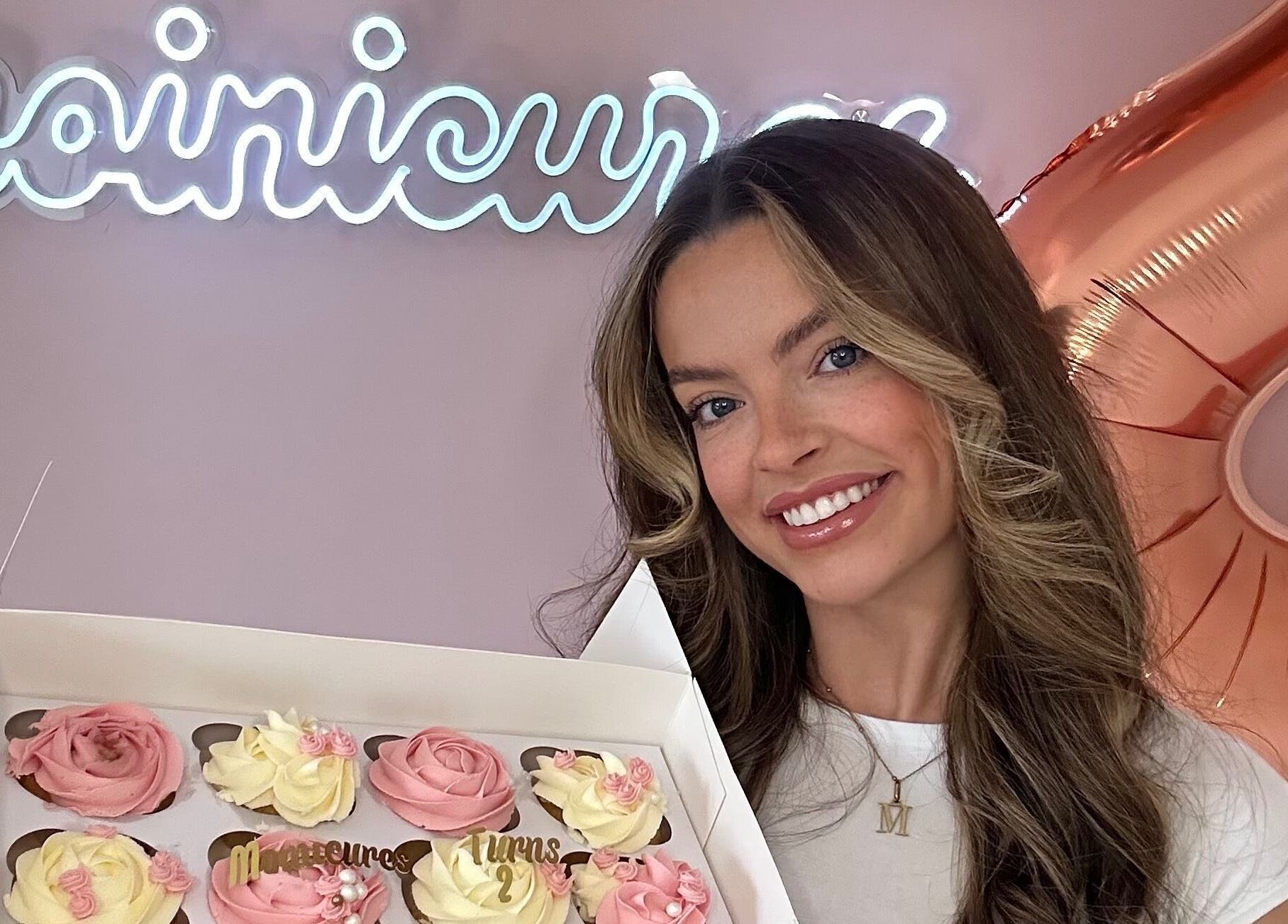 Smiling woman holds cupcakes at Moinicures, Barry, Wales, GB with venue sign and balloons in the background.
