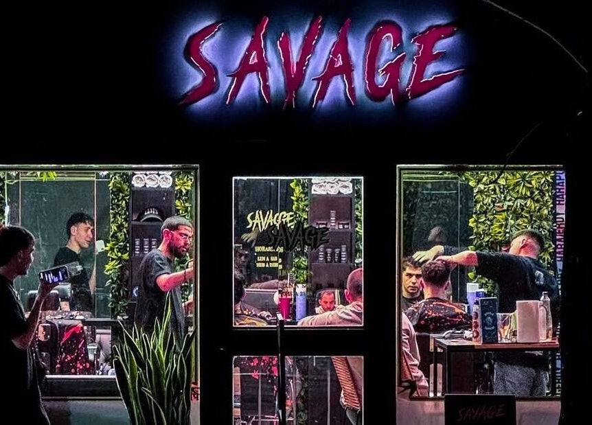 Vista nocturna de Savage Barber Club en Buenos Aires, con barberos en acción en un ambiente moderno y vibrante.
