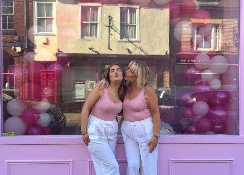 Two women in matching outfits embrace in front of Lotties Lashes, Sudbury, England, GB, with festive balloons.