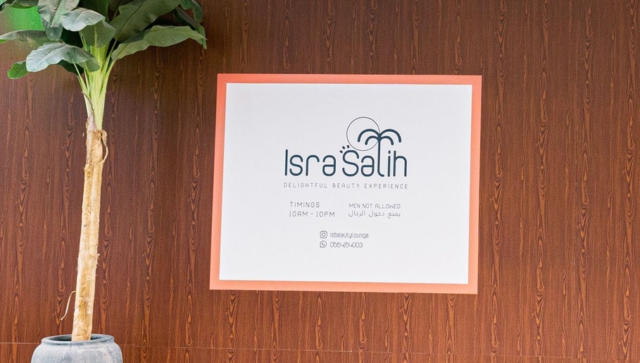 Welcome sign at Isra Salih Beauty Lounge, Sharjah, with timings and contact info on a wooden wall background.