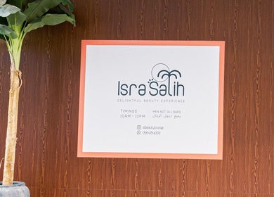 Welcome sign at Isra Salih Beauty Lounge, Sharjah, with timings and contact info on a wooden wall background.