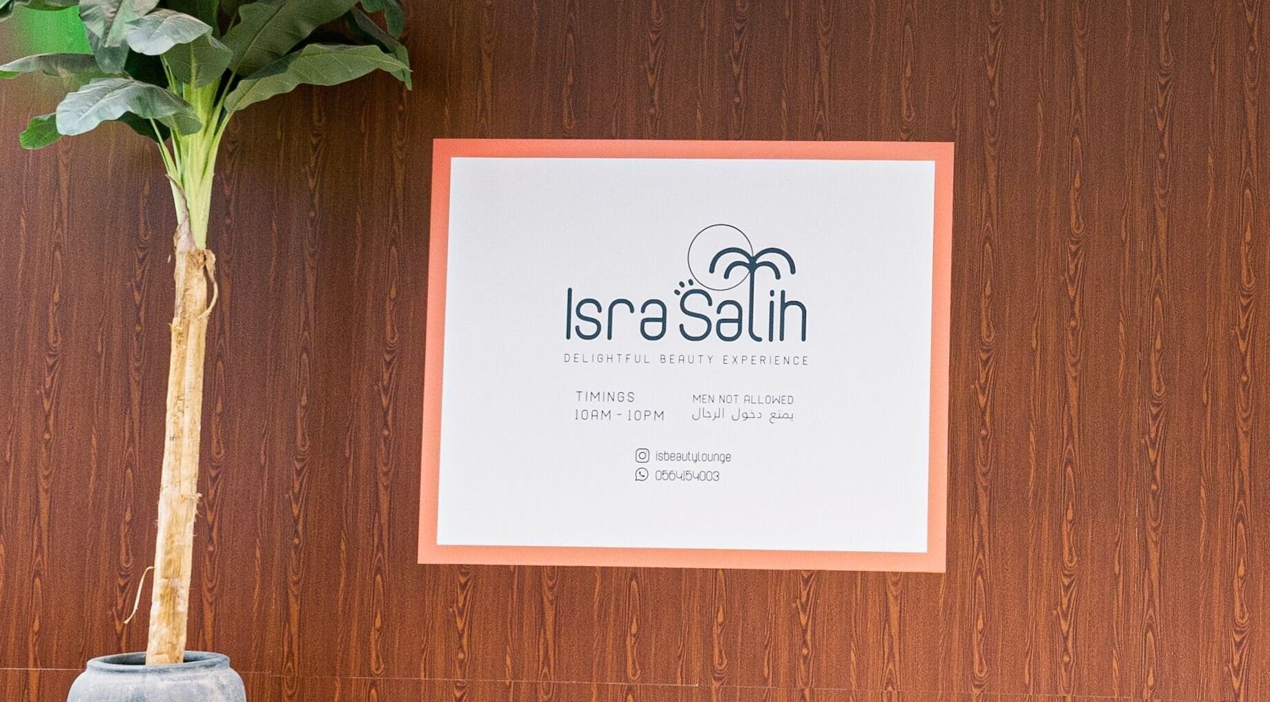 Welcome sign at Isra Salih Beauty Lounge, Sharjah, with timings and contact info on a wooden wall background.
