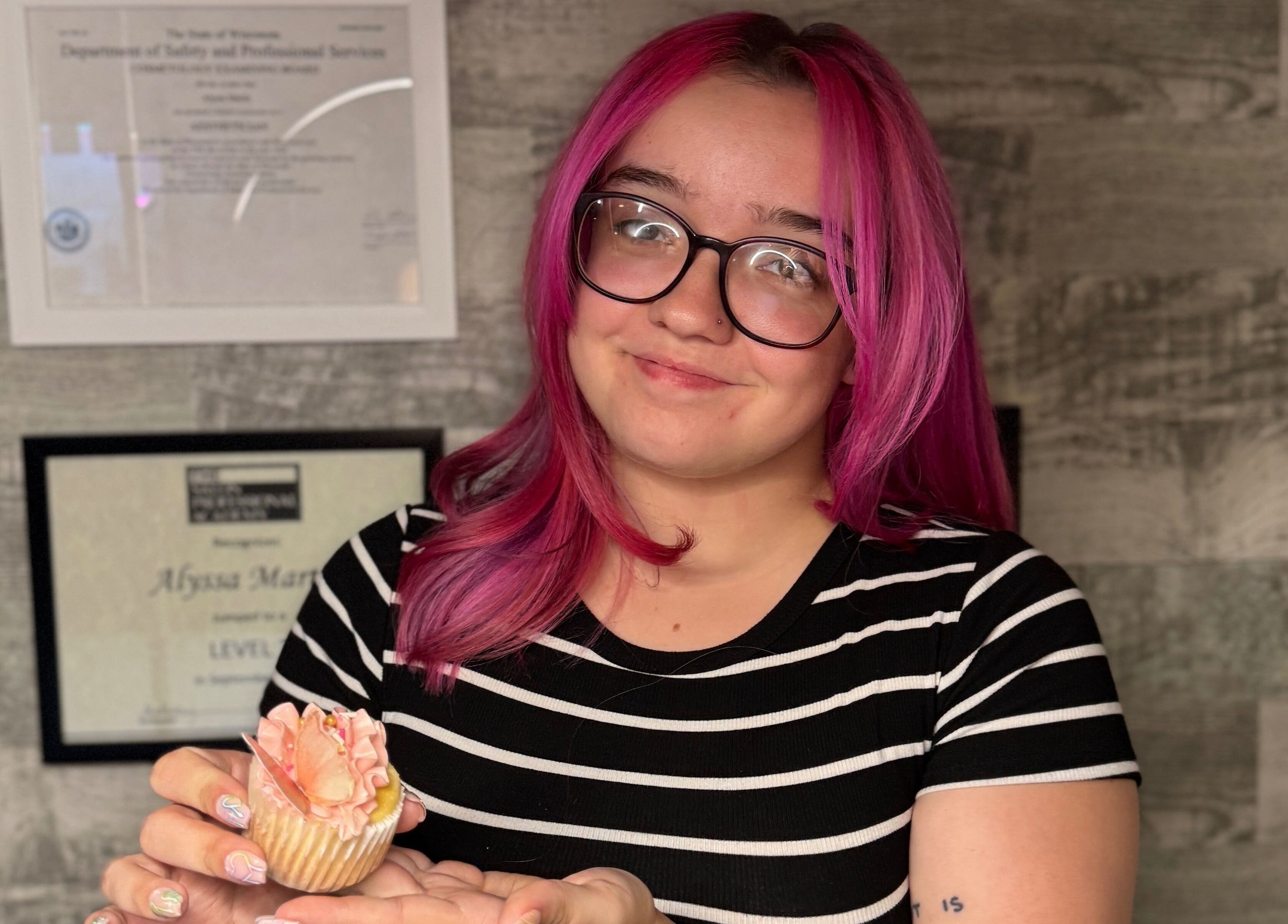 Person with pink hair holding a cupcake at Timeless Glow Aesthetics, Kenosha, Wisconsin, US.