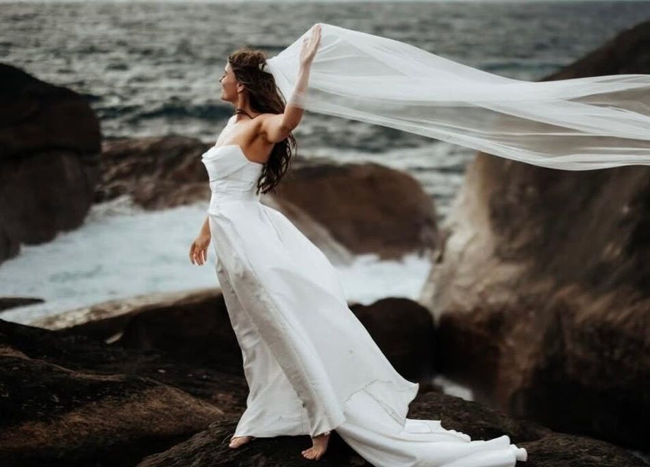 Bride in flowing gown by ocean rocks, captured at Bronzed Body Spray Tan Studio, Port Kennedy, Western Australia, AU.