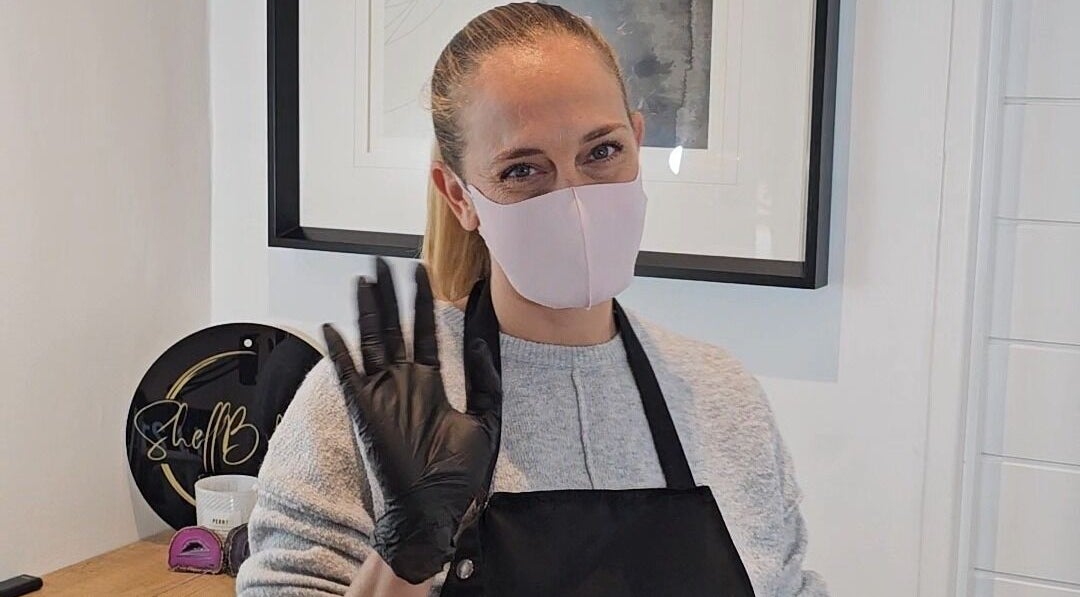 Staff member at ShellB Nails, Cape Town, Western Cape, ZA, greets with a warm gesture while wearing a mask and gloves.