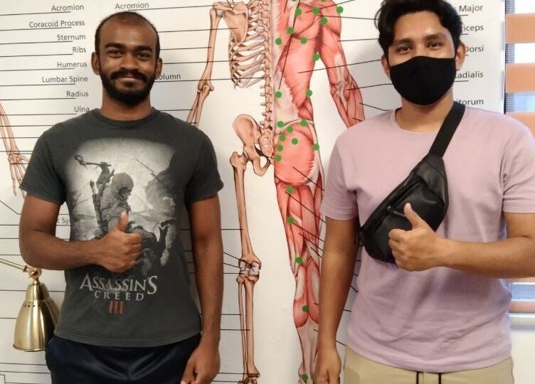 Two clients smiling at Advanced Muscle Therapy in Petaling Jaya, Selangor, MY, thumbs up near anatomy chart.