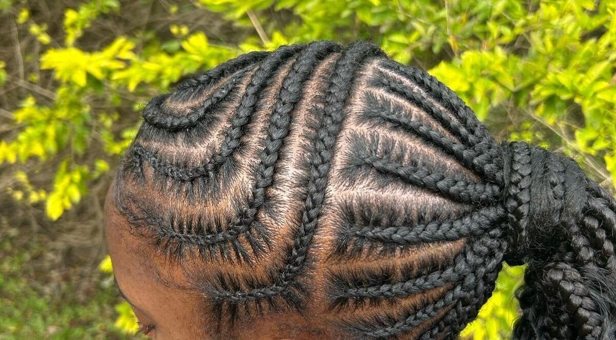 Intricate cornrow braids hairstyle at Harmonized Hairstyles, Babonneau, LC against green foliage background.