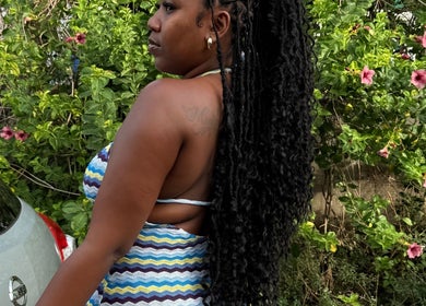 Woman with long braided hair at SlayedbyKy in Brighton, St. George, VC, surrounded by vibrant foliage.