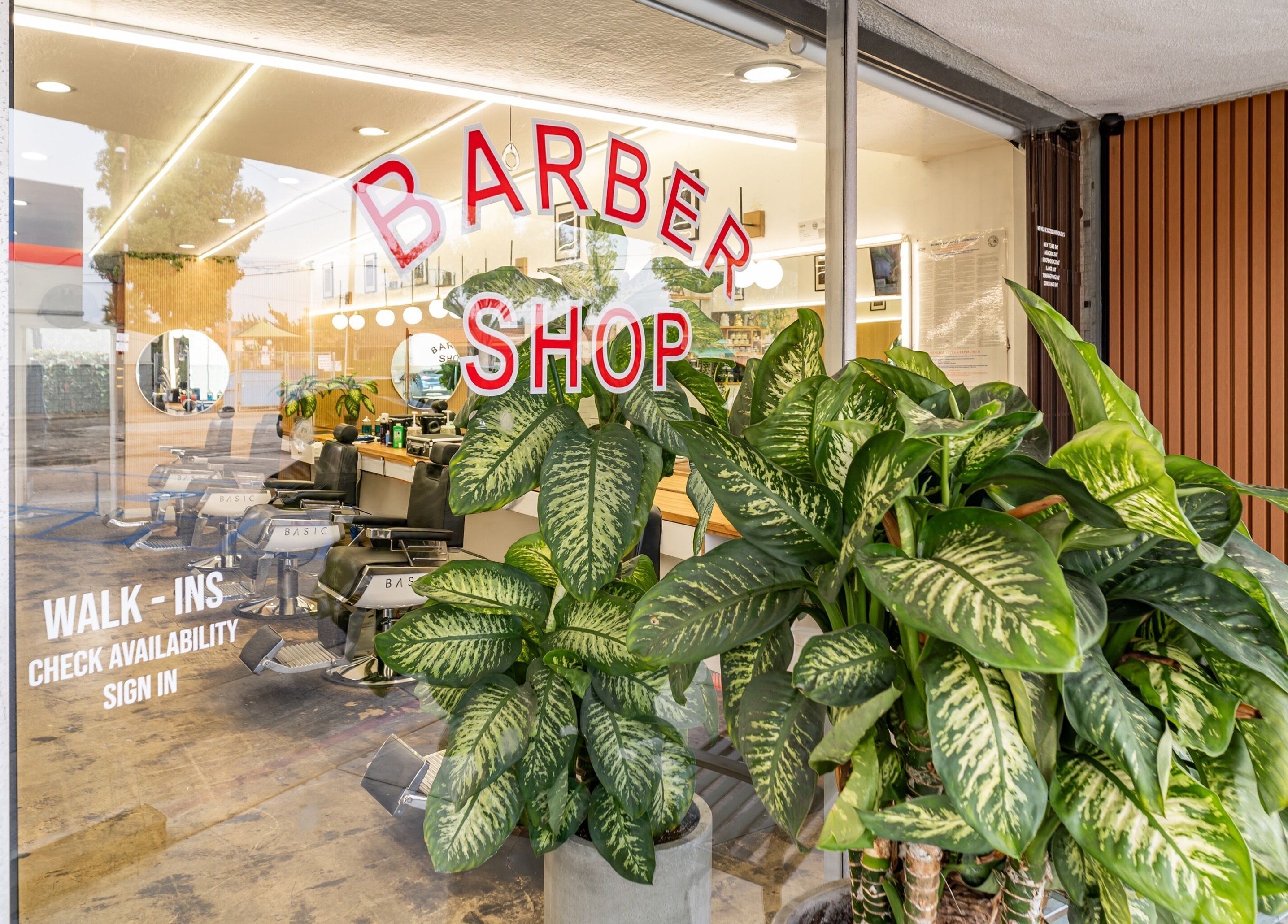 Stylish entrance of Basic Barber in Costa Mesa, California, US with vibrant plants and barber chairs visible inside.