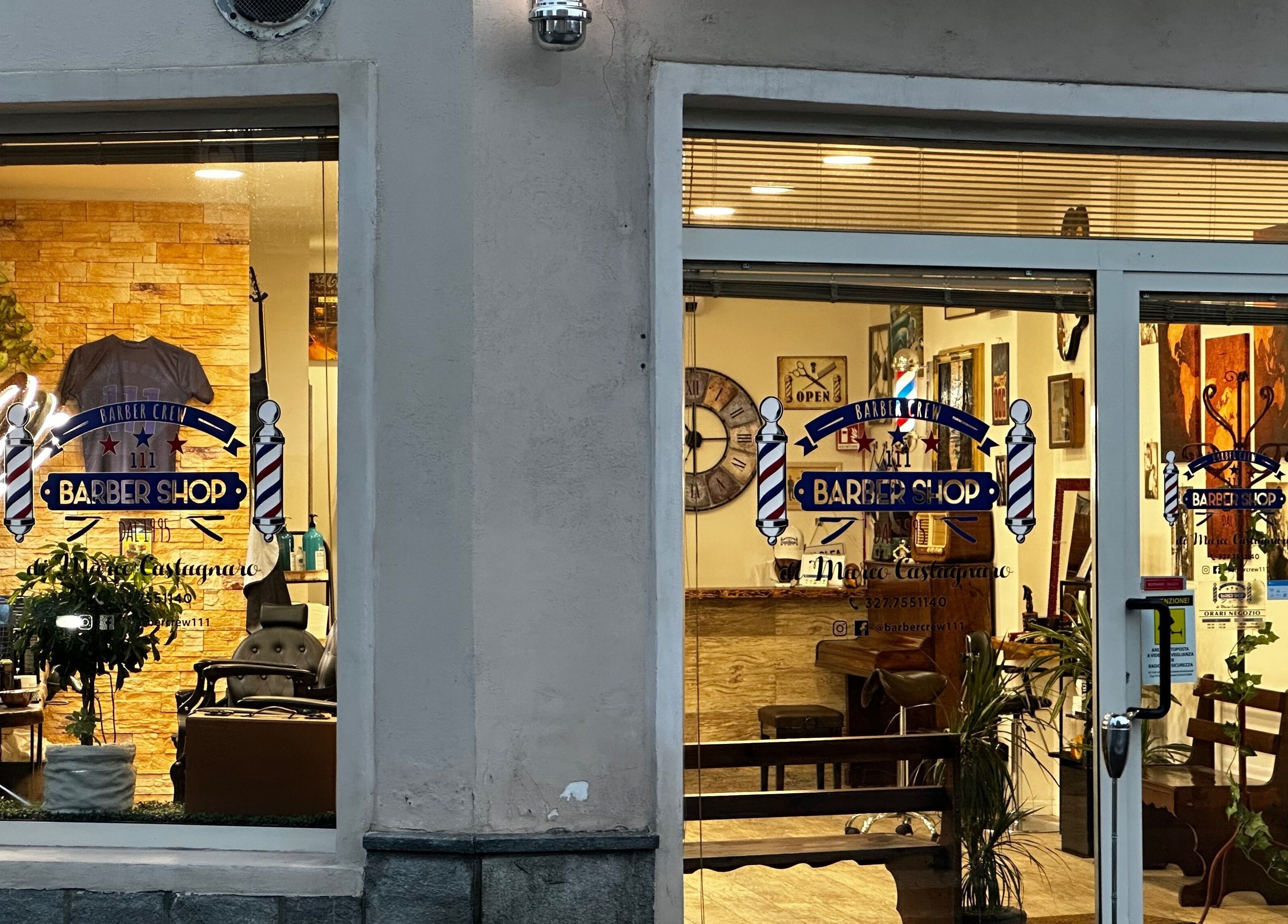 Vetrina Barber Crew 111, a Asti, Piemonte, IT, illuminata di sera con sedie vintage all'interno.