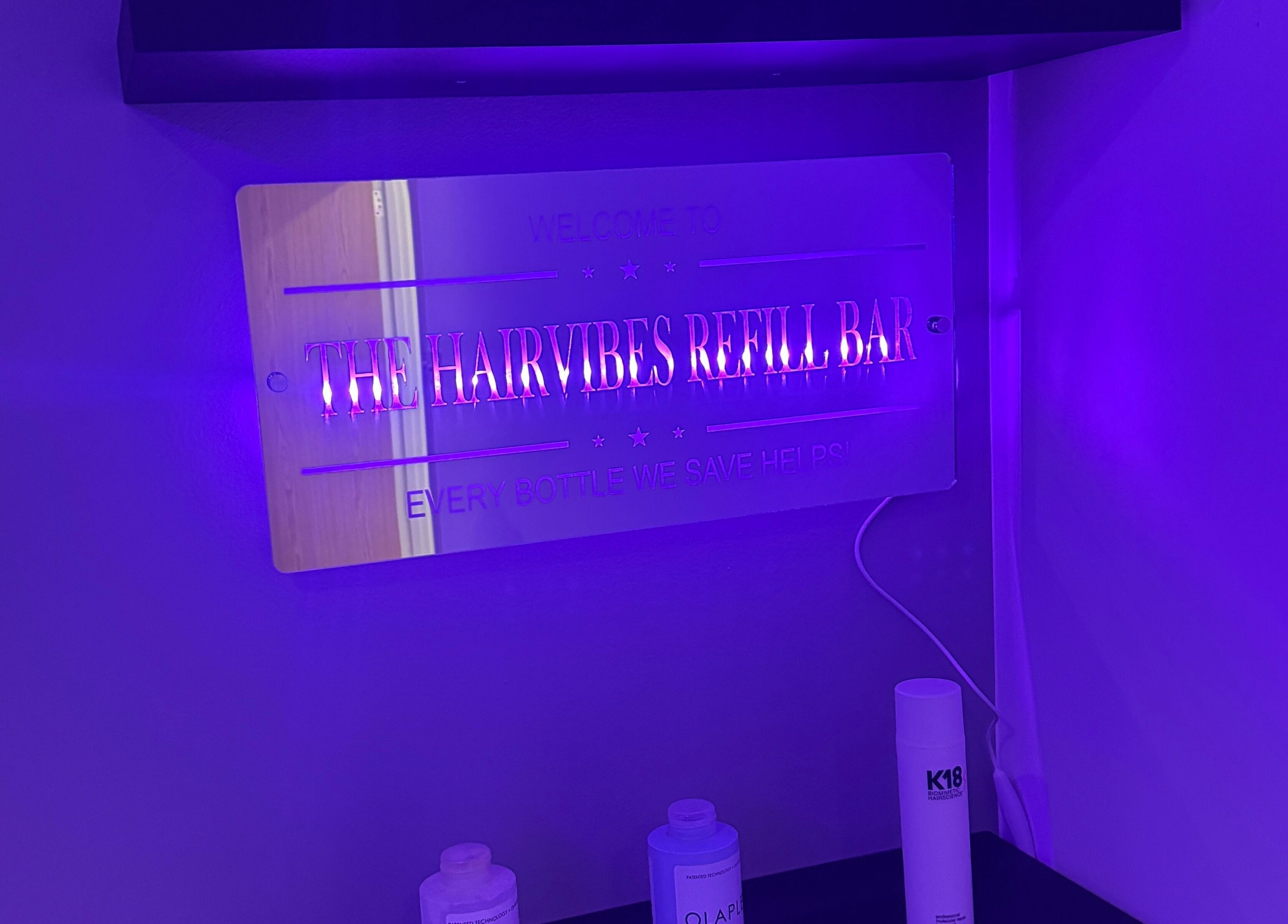 Illuminated Hairvibes refill bar sign with products in Hazlemere, England, GB, under purple lighting.