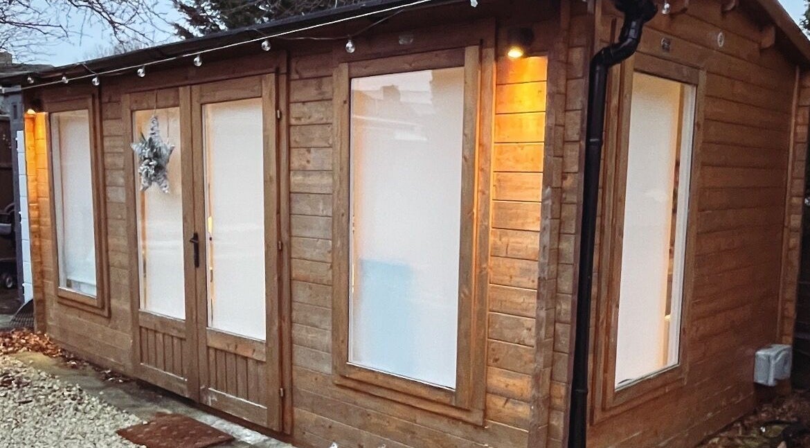 Rustic wooden exterior of Jayme’s Aesthetics in Hornchurch, England, GB, warmly lit at dusk.