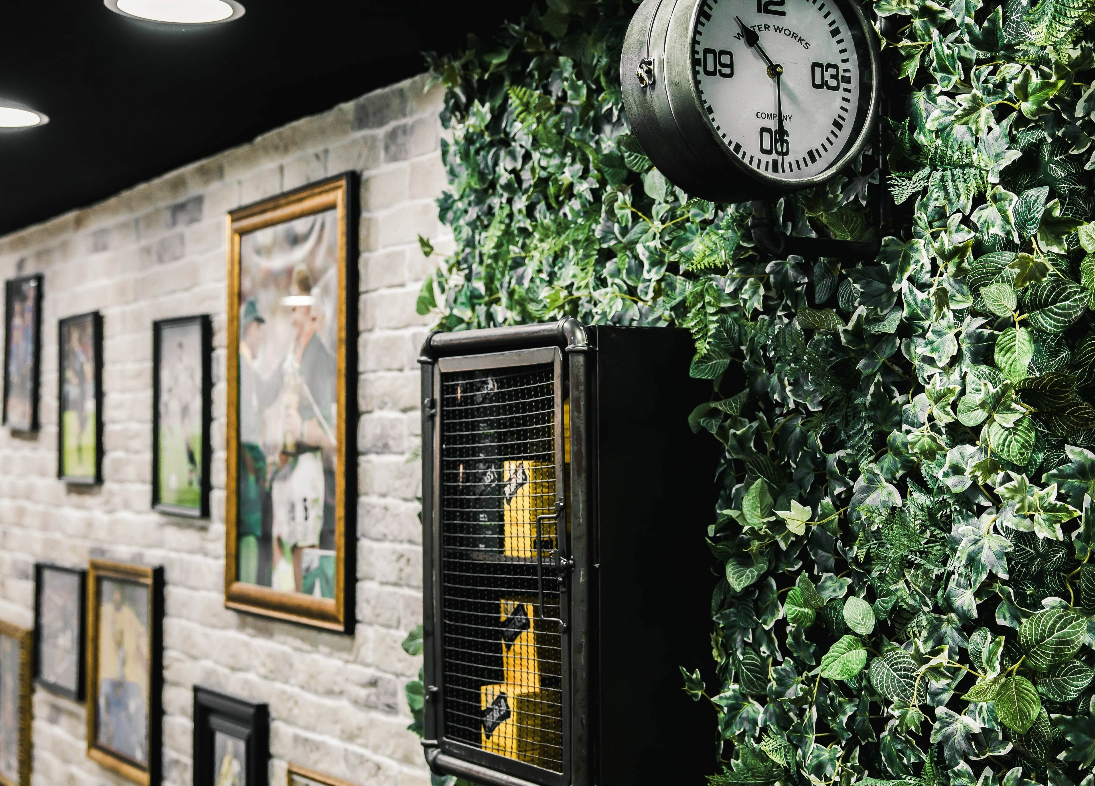 Eclectic interior at Strak Strak - Barbershop, Rotterdam, with framed art and a vintage clock on green foliage wall.