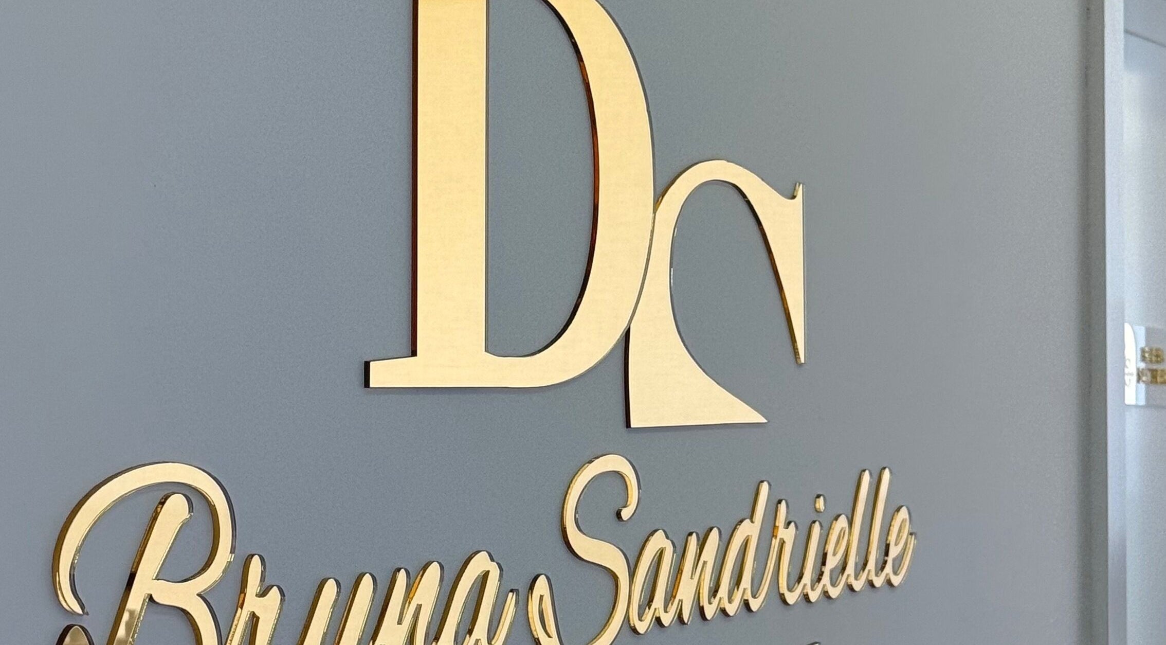 Logotipo dourado da Sandrielle beauty concept em Padrão Da Légua, Porto, PT em uma parede cinza.
