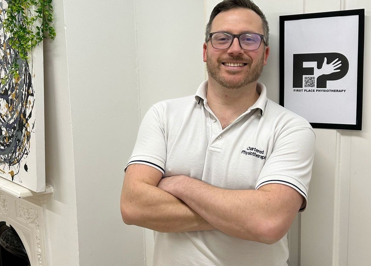 Therapist at First Place Physiotherapy in Fleet, England, GB, standing in front of art and the clinic logo.