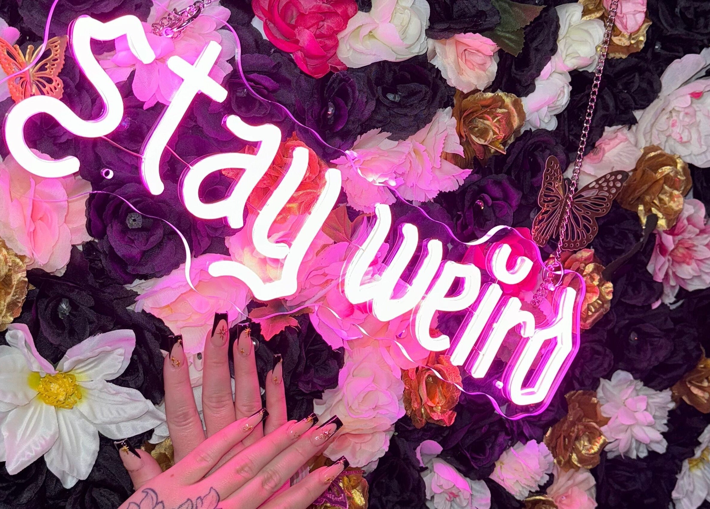Floral backdrop with neon 'stay weird' sign and manicured hand at Beauty Girl Studios, Toronto, Ontario, CA.