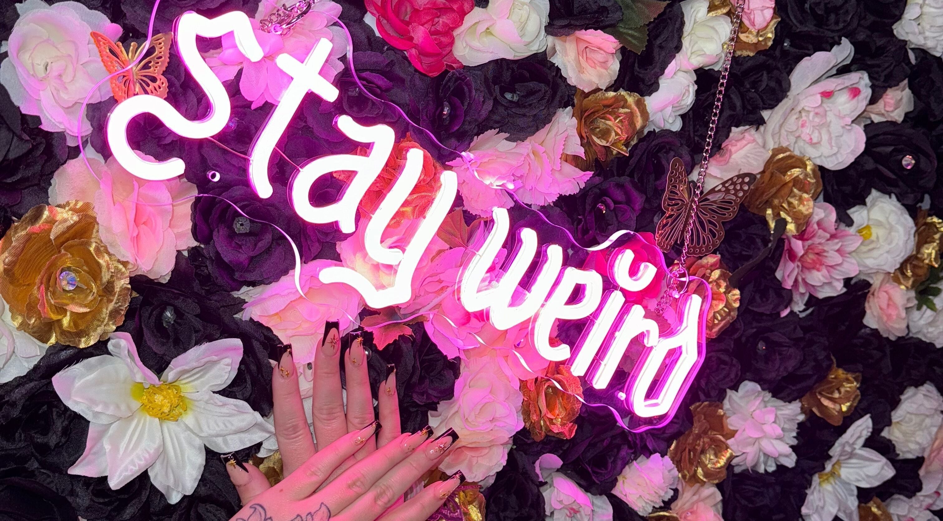 Floral backdrop with neon 'stay weird' sign and manicured hand at Beauty Girl Studios, Toronto, Ontario, CA.