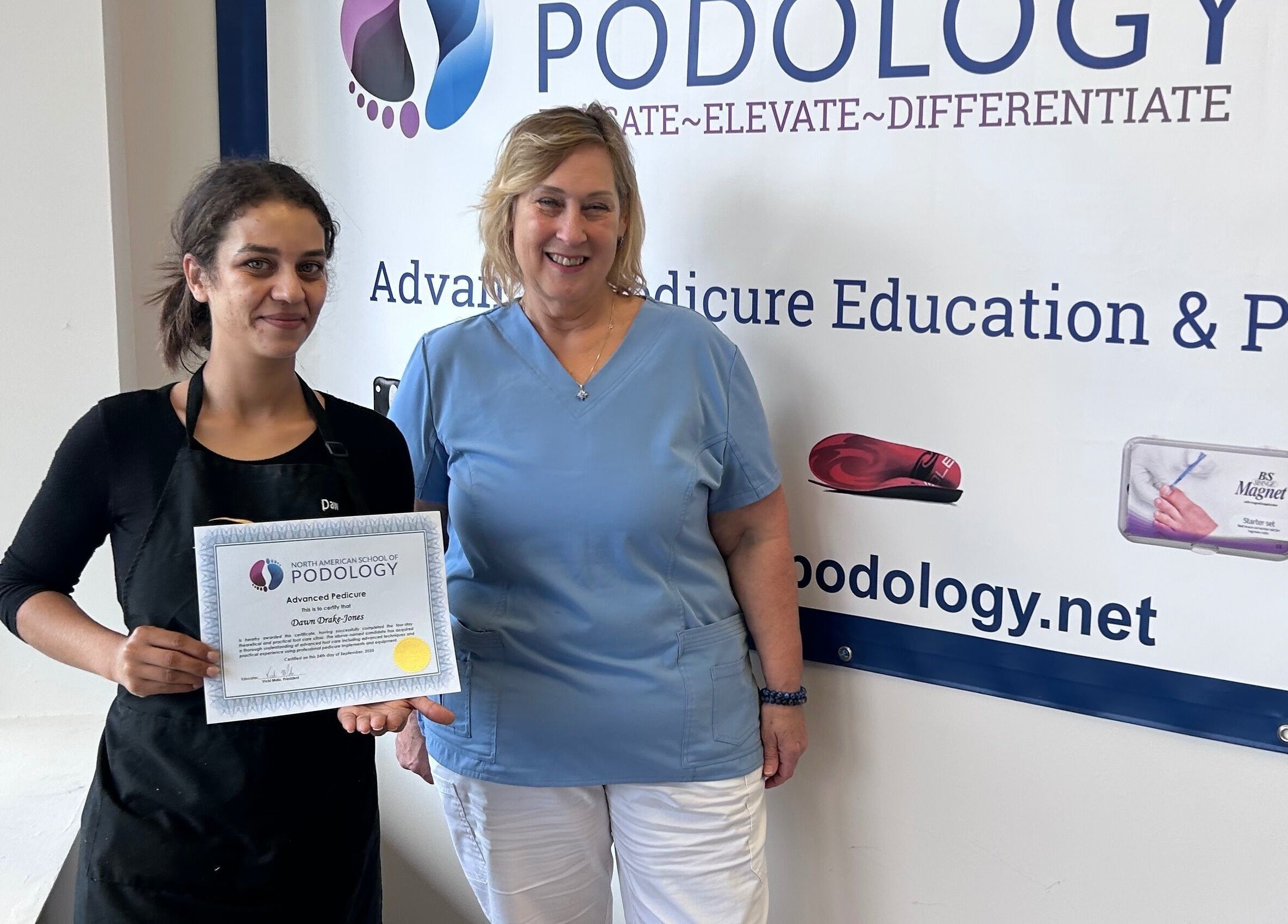 Two women at Beauty Girl Studios in Toronto hold a podology certificate, smiling near a large logo backdrop.