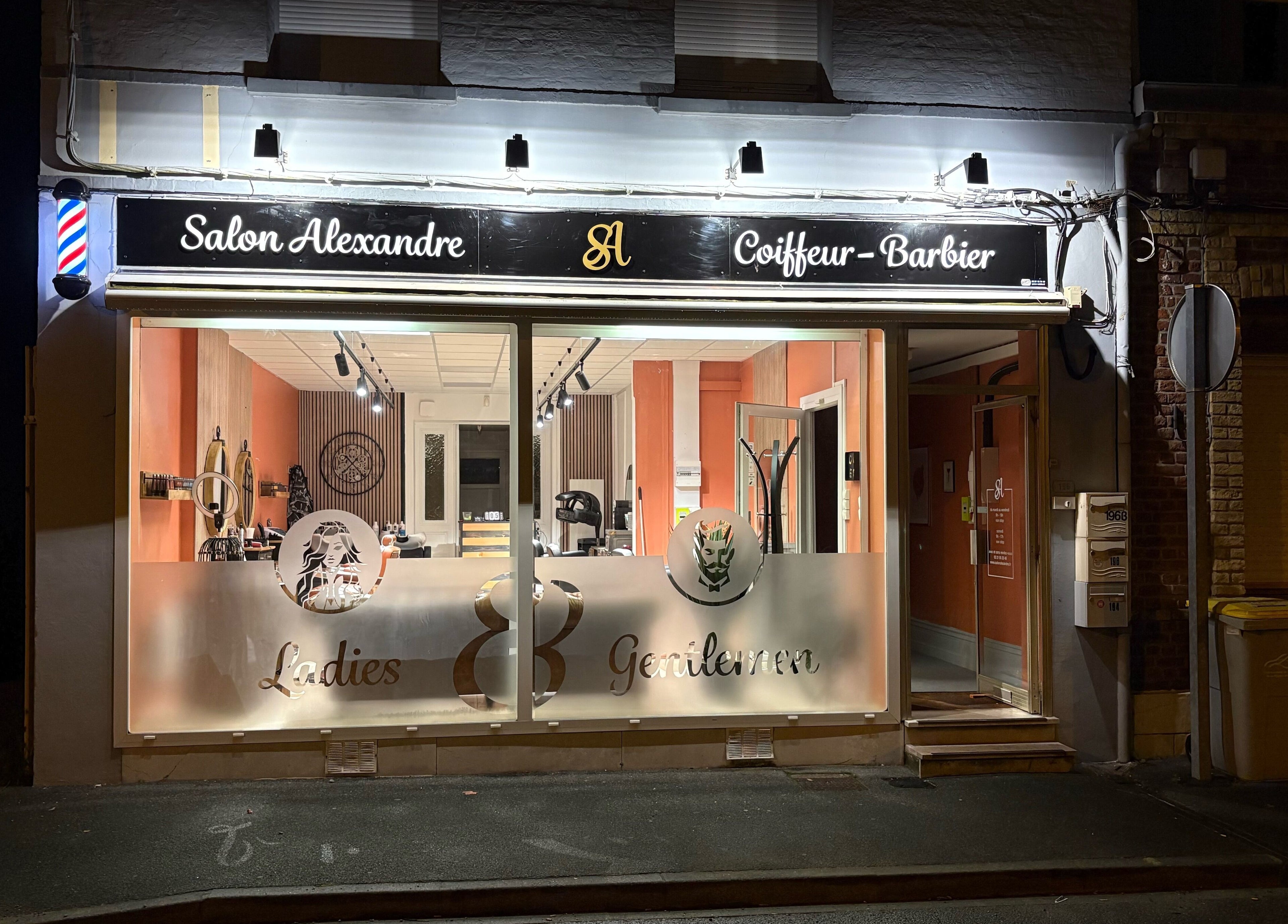 Façade de Salon Alexandre à Audruicq, Hauts-de-France, FR, éclairée la nuit, affichant le logo et la devanture.