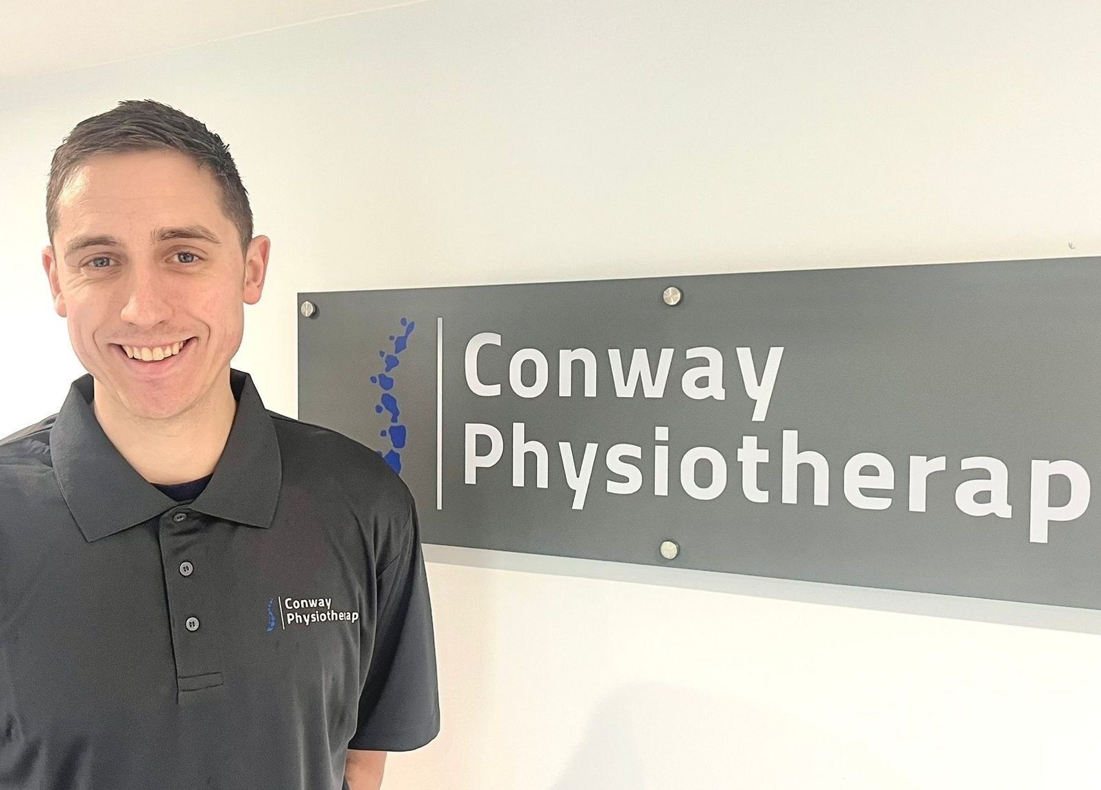 Smiling therapist at Conway Physiotherapy, Ratby, England, GB beside venue sign.