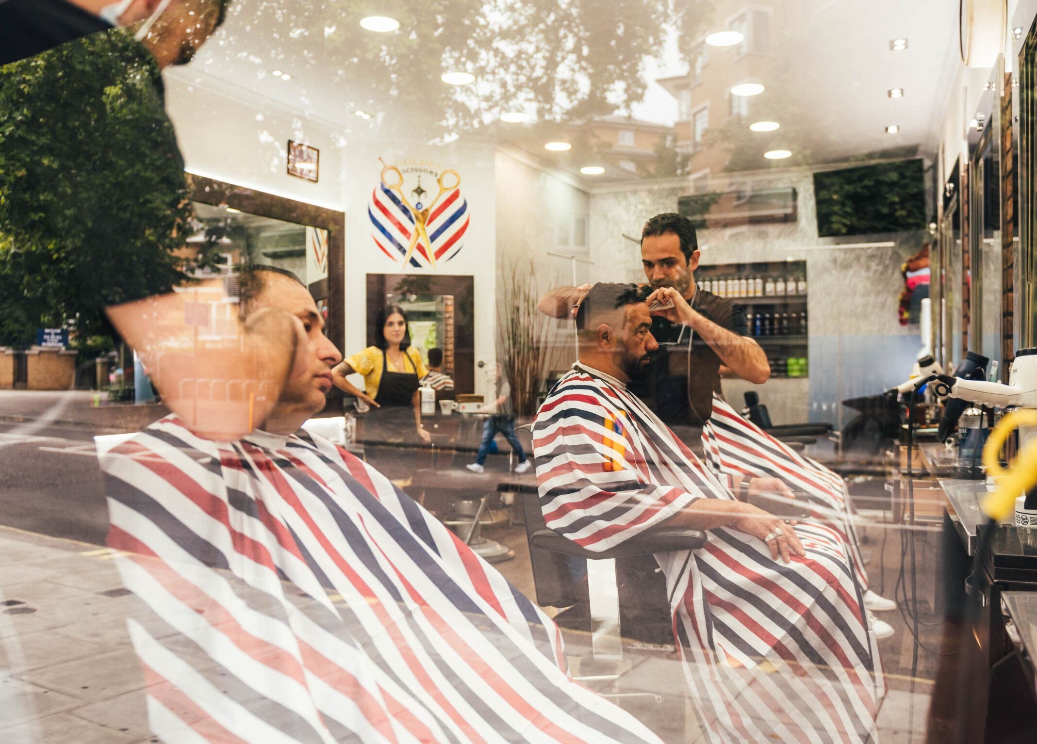 Inside view of GOLDEN SCISSORS barbershop, London, England. Clients in striped capes, stylish trims by skilled barber.
