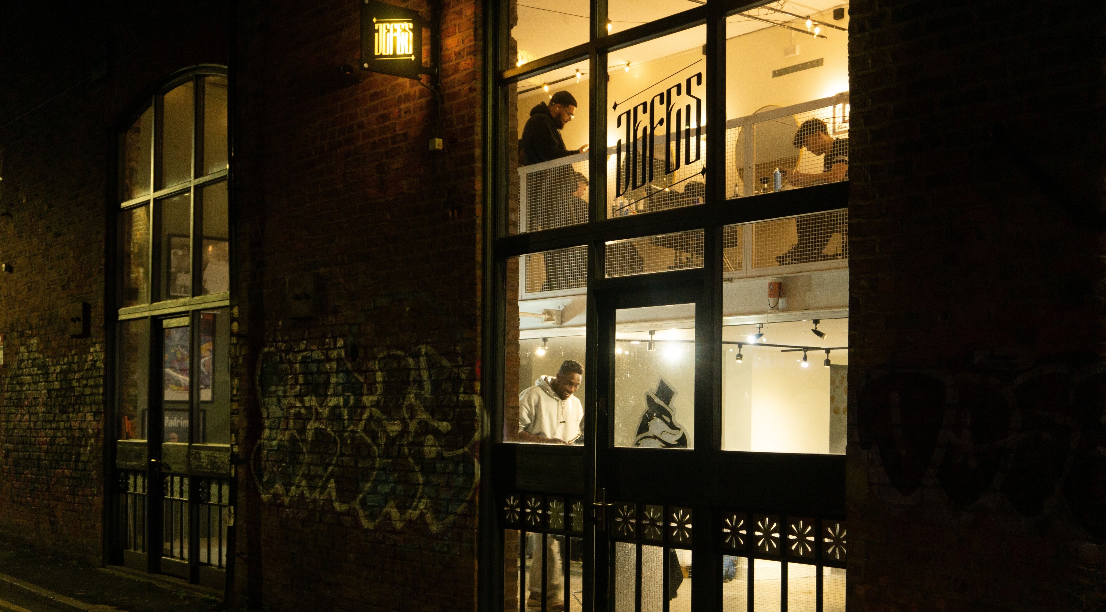 Exterior of Jefe's Barbers in Manchester, England, GB lit warmly at night, showcasing vibrant street scene.