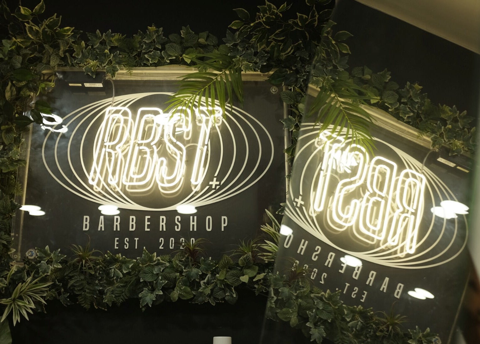 Illuminated RBST+ Barber Shop neon sign with lush greenery in Horwich, England, GB reflecting vibrant ambiance.