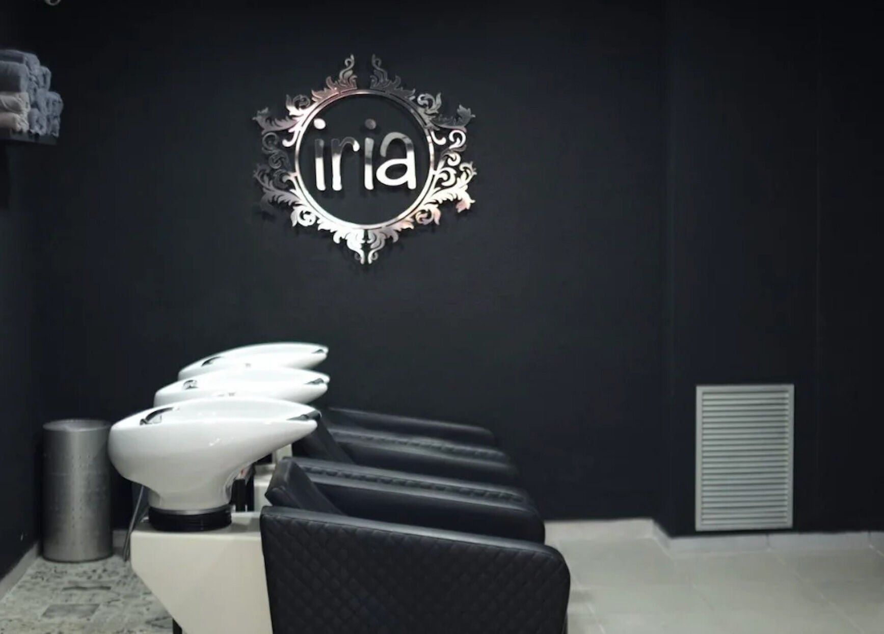 Zona de lavado en Iria Perruquers, Cubelles, Catalunya, ES con sillas negras y un elegante logo de pared.