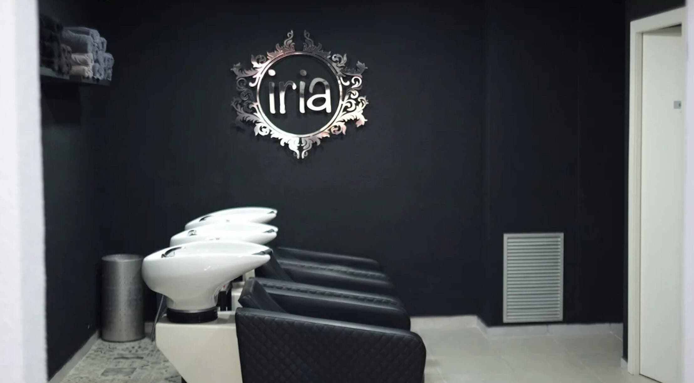 Zona de lavado en Iria Perruquers, Cubelles, Catalunya, ES con sillas negras y un elegante logo de pared.