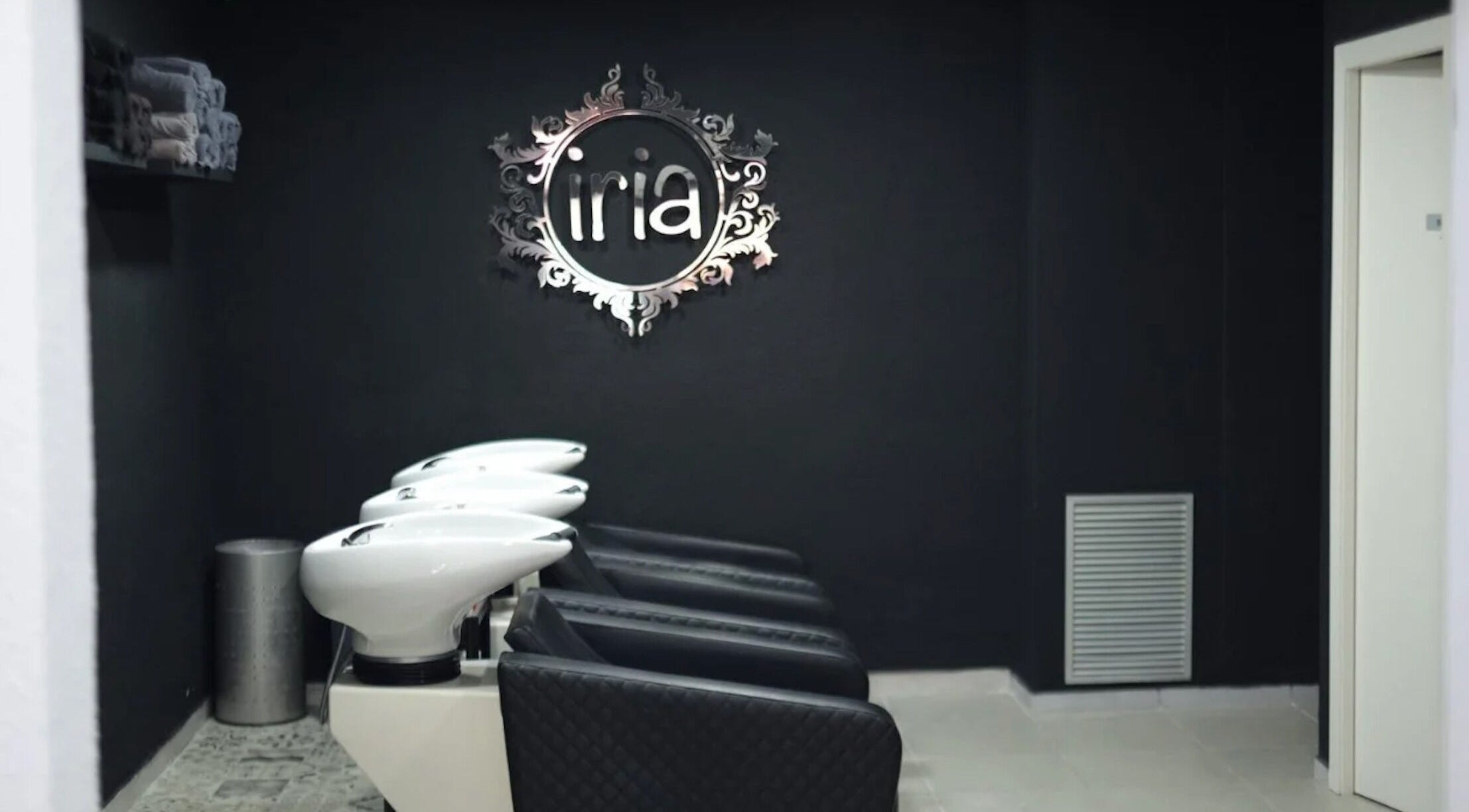 Zona de lavado en Iria Perruquers, Cubelles, Catalunya, ES con sillas negras y un elegante logo de pared.