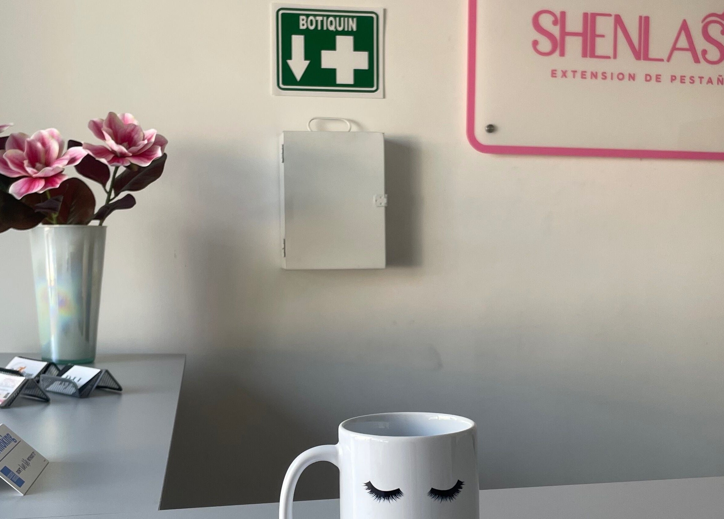 Interior de Shenlash en Santiago De Querétaro, MX, con taza decorativa, flores y señal de botiquín.