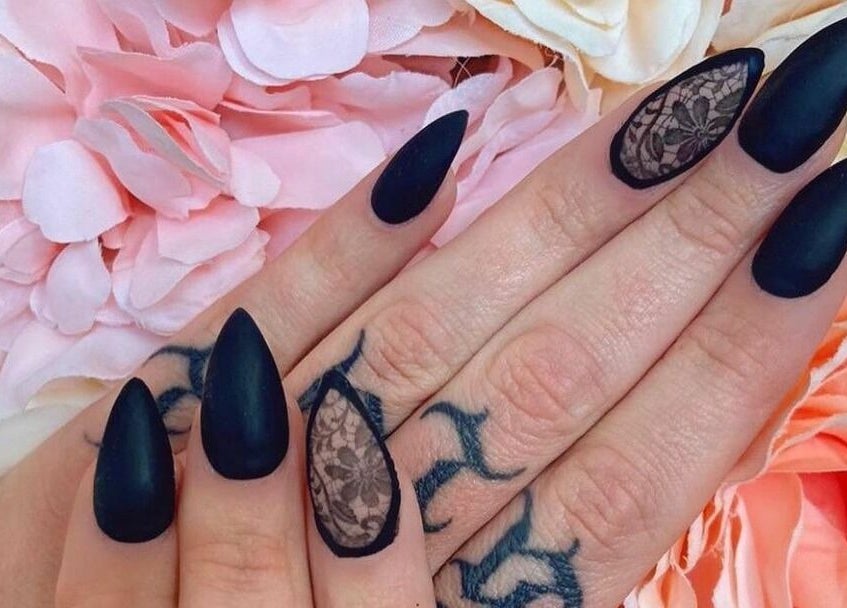 Black matte stiletto nails with lace design at Polished, Torquay, England, GB, surrounded by pink flowers.
