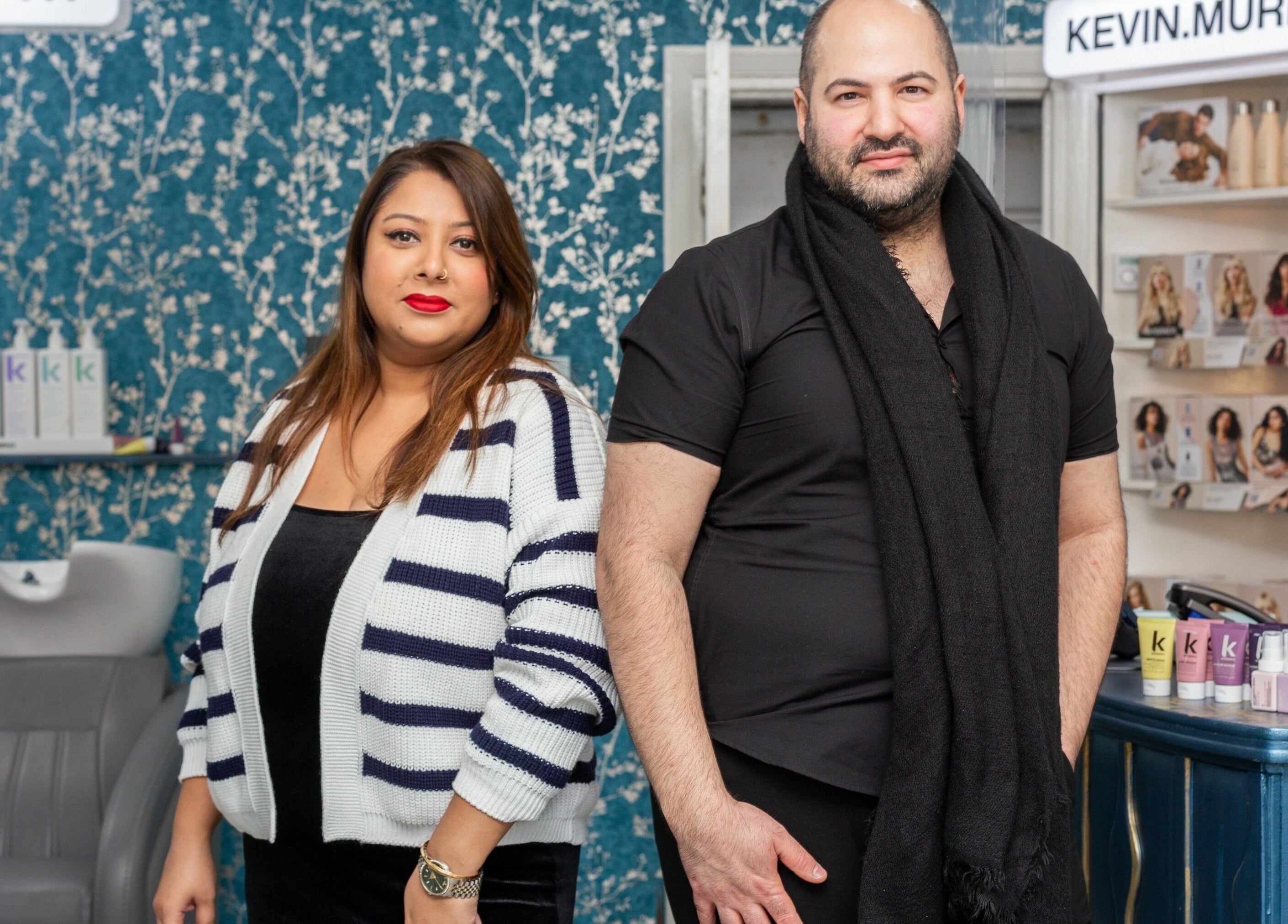 Two staff members at The Beauty Inn, London, England, GB, standing in front of elegant wallpaper and beauty products.