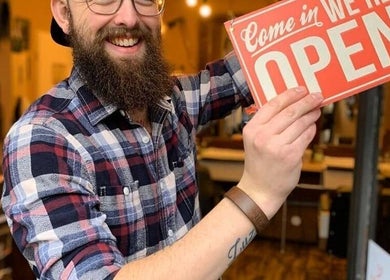 Smiling staff at The Canadian Barbershop in Praha, CZ holding an open sign inside the cozy shop.