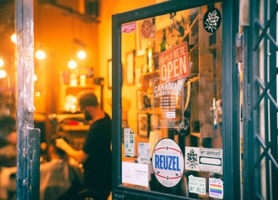 Warm interior view of The Canadian Barbershop, Praha, CZ, through a glass door with welcoming signs.