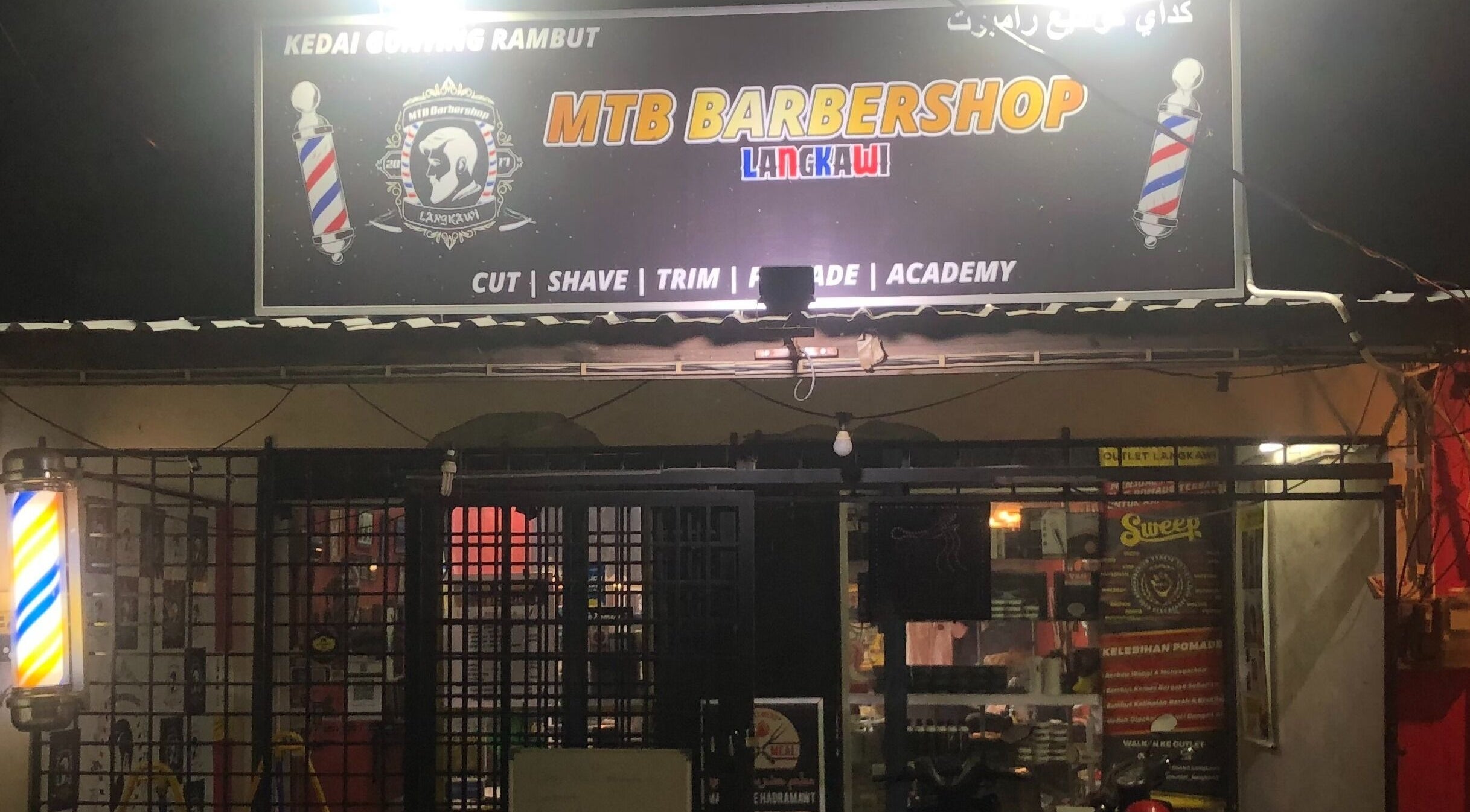 MTB Barbershop and Academy in Langkawi, Kedah, MY, lit up at night showcasing its vibrant sign and entrance.