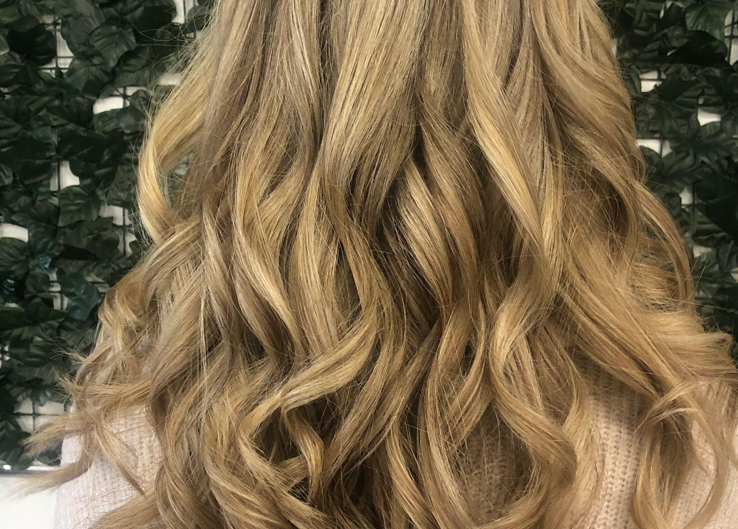Long wavy blonde hair styled at Hair and Body, Colchester, England, GB against a lush green wall.