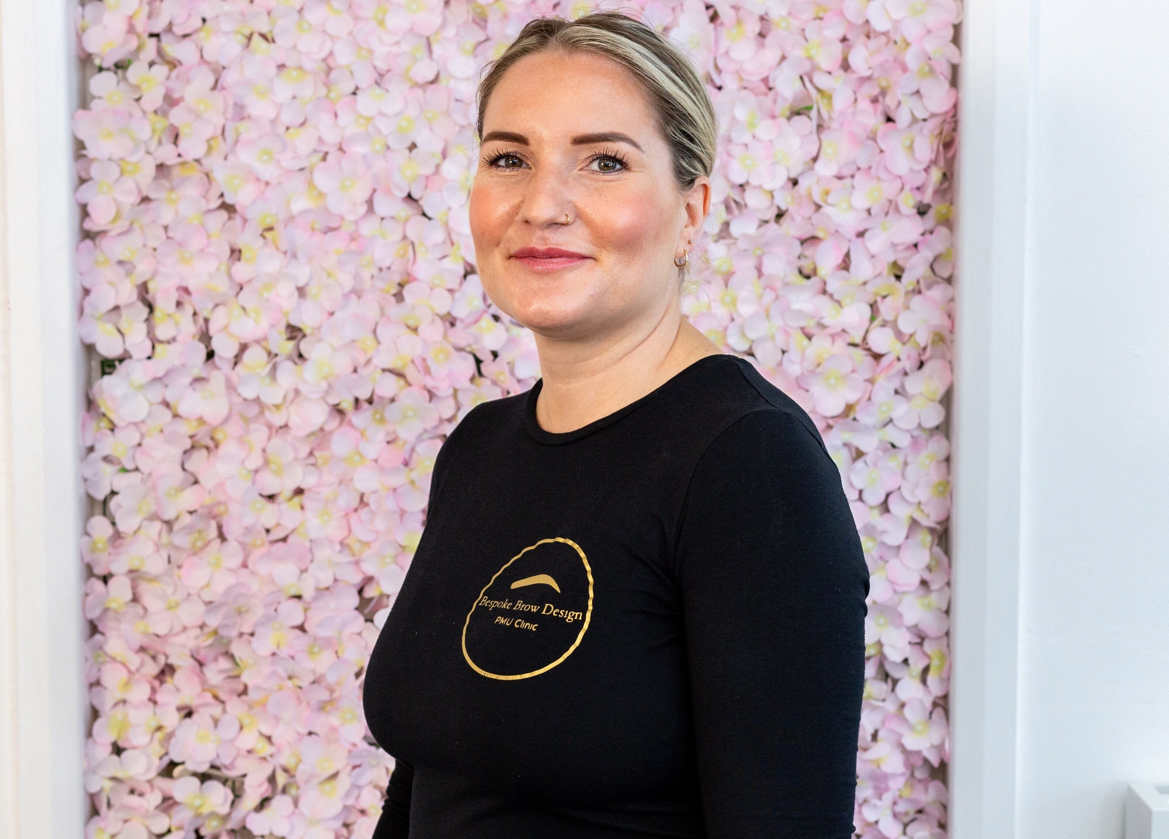 A professional at Bespoke Brow Design, Rustington, England, GB, standing before a pink floral backdrop.