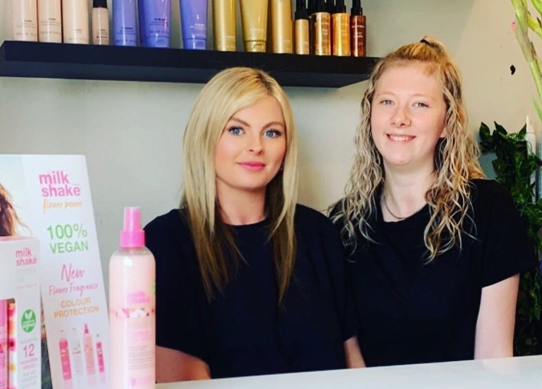 Two smiling stylists at the reception desk of Salon Seven, Wivenhoe, England, GB, with beauty products displayed.