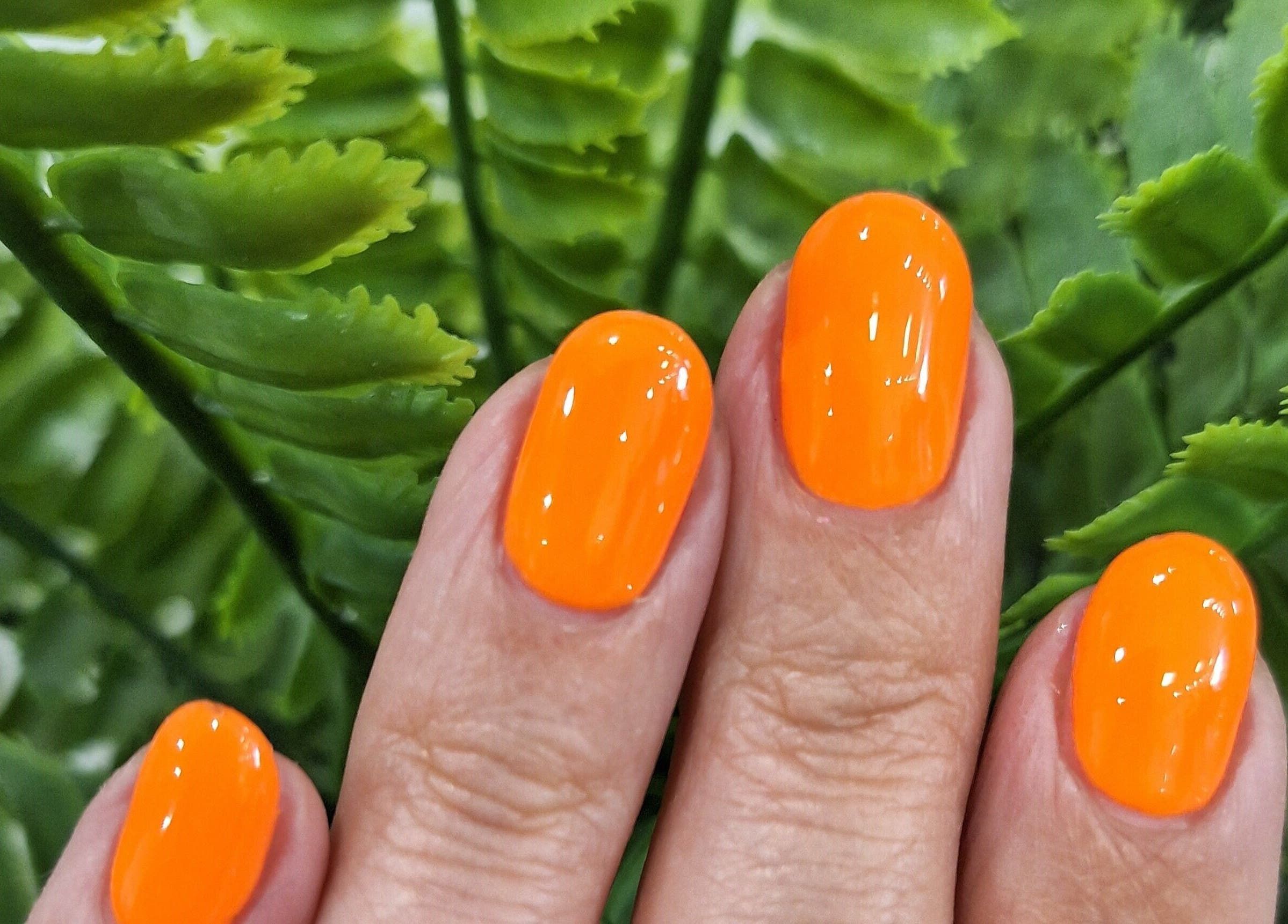 Bright orange nail polish on manicured nails at Xar Hair & Nails in Is-Swieqi, MT amid lush green leaves.