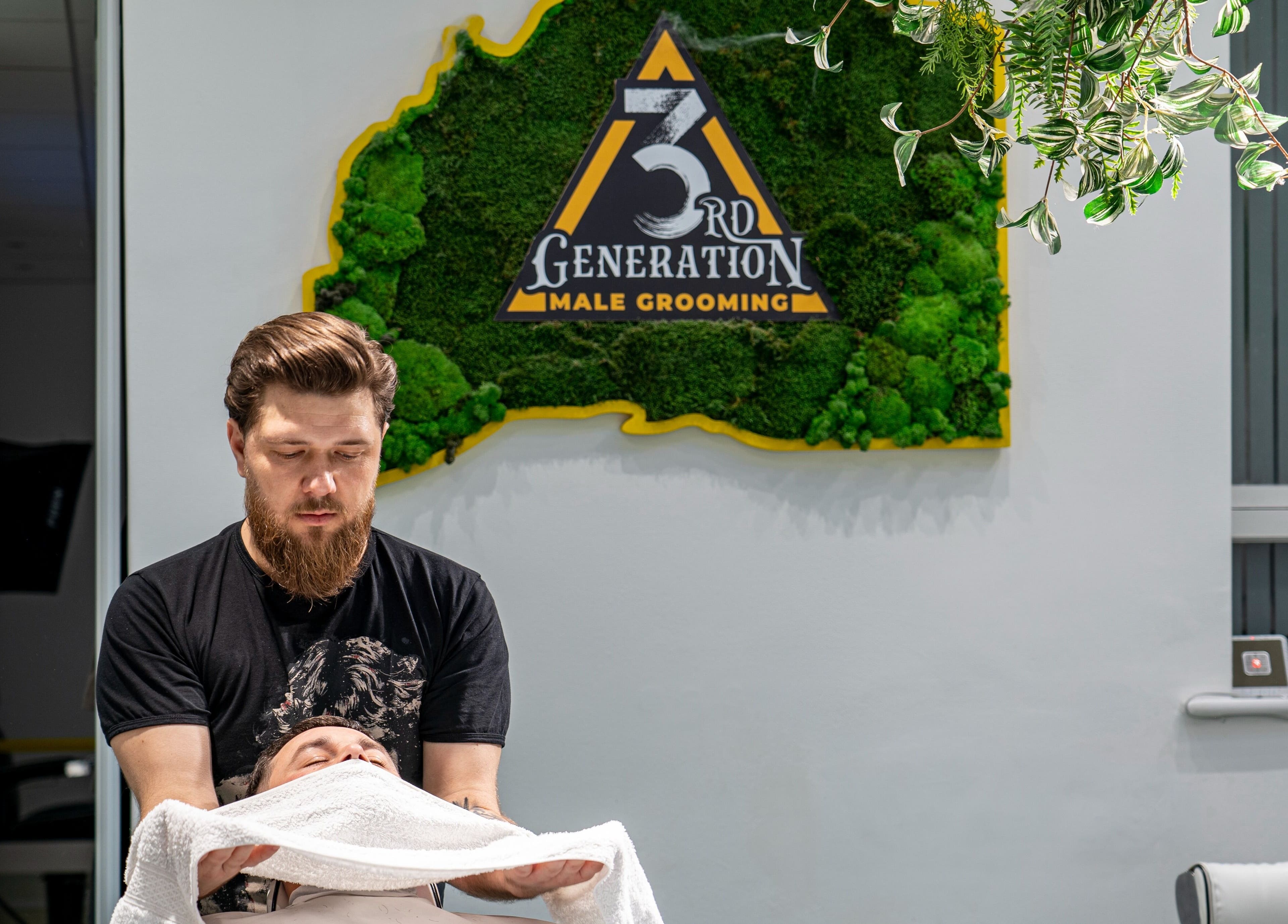 Barber performs grooming at 3rd Generation Grooming in London, England, GB, under a lush green wall sign.