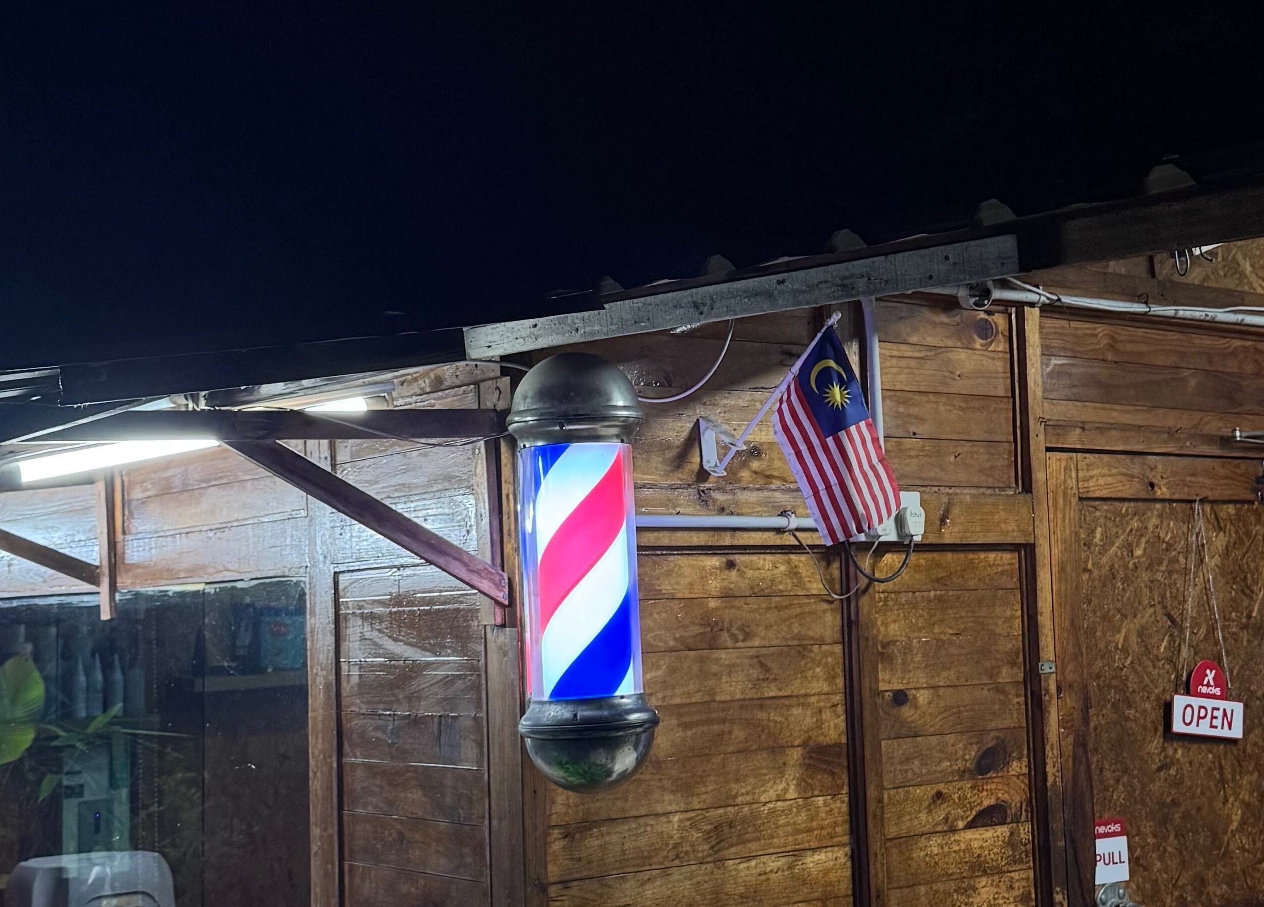 Aididthebarber in Klang, Selangor, MY featuring a lit barber pole at night next to Malaysian flag.